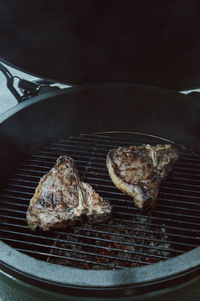 Twee gegrilde steaks op de bbq van een Green Egg, met houtskool dat gloeit onder het rooster.