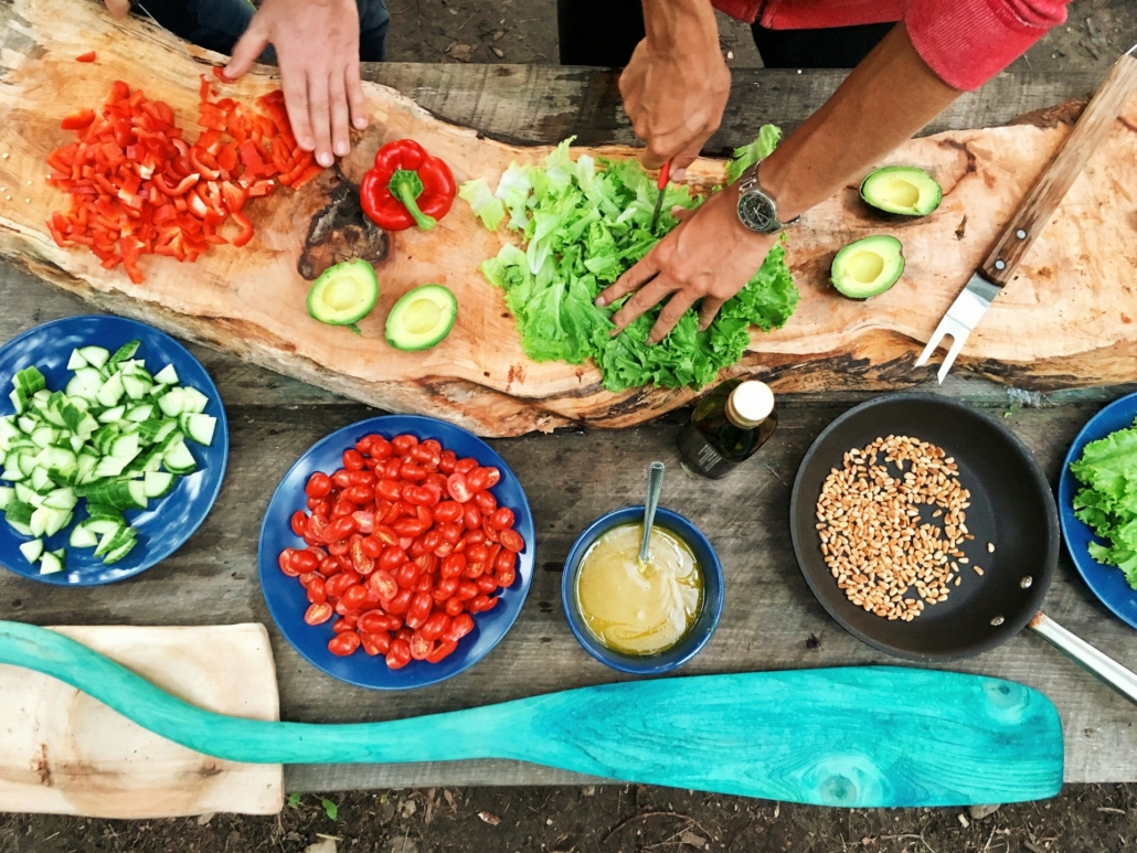 Mensen bereiden verse groenten voor op een rustiek houten plank tijdens het buiten koken, met ingrediënten zoals paprika, avocado, tomaten en komkommer. Verschillende gerechten en zaden liggen verspreid op de tafel.