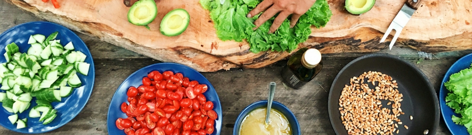 Mensen bereiden verse groenten voor op een rustiek houten plank tijdens het buiten koken, met ingrediënten zoals paprika, avocado, tomaten en komkommer. Verschillende gerechten en zaden liggen verspreid op de tafel.