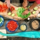 Mensen bereiden verse groenten voor op een rustiek houten plank tijdens het buiten koken, met ingrediënten zoals paprika, avocado, tomaten en komkommer. Verschillende gerechten en zaden liggen verspreid op de tafel.