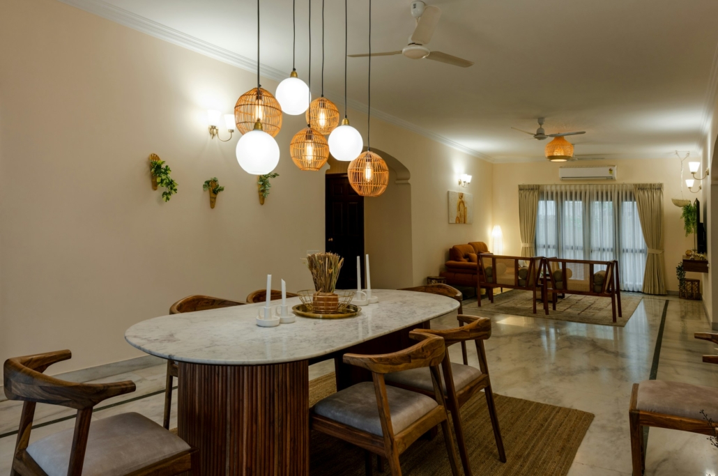 Eetkamer met een ovale tafel van marmer, omringd door houten stoelen met grijze kussens, hangende rieten en glazen lampen erboven, en een lichte woonkamer op de achtergrond.