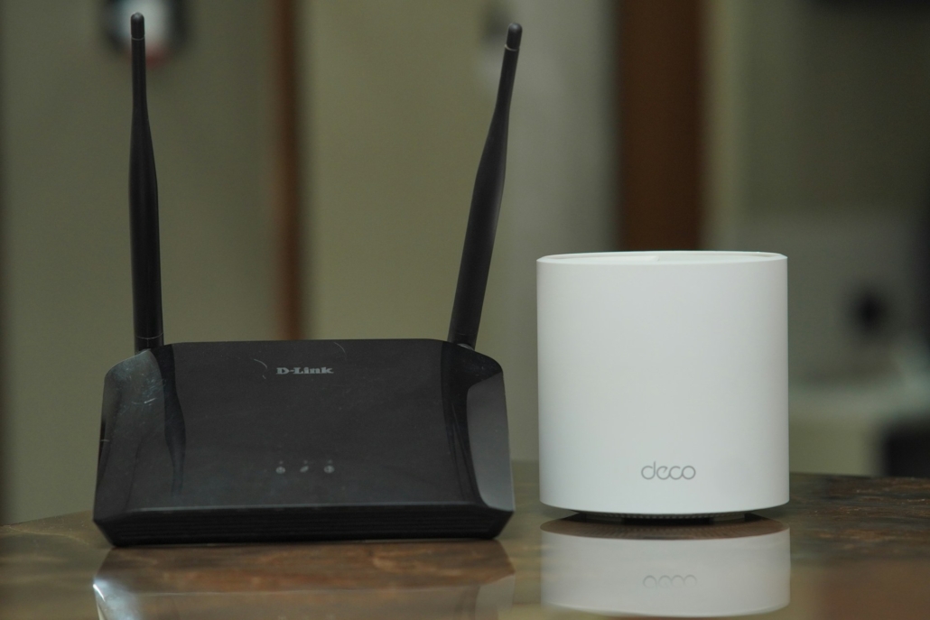 Zwarte D-Link router naast een witte Deco mesh Wi-Fi-unit, geplaatst op een tafel. Dit toont een vergelijking tussen traditionele routers en moderne mesh-netwerkoplossingen.