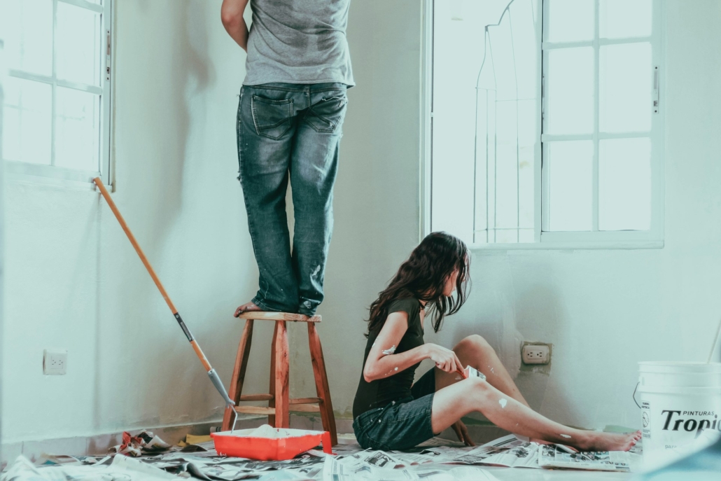 Man en vrouw bezig met verbouwingen, schilderen van een muur in een kamer. De man staat op een kruk terwijl de vrouw op de grond zit met een verfkwast in haar hand, omringd door verfbenodigdheden.