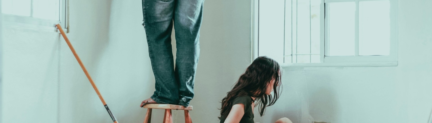 Man en vrouw bezig met verbouwingen, schilderen van een muur in een kamer. De man staat op een kruk terwijl de vrouw op de grond zit met een verfkwast in haar hand, omringd door verfbenodigdheden.