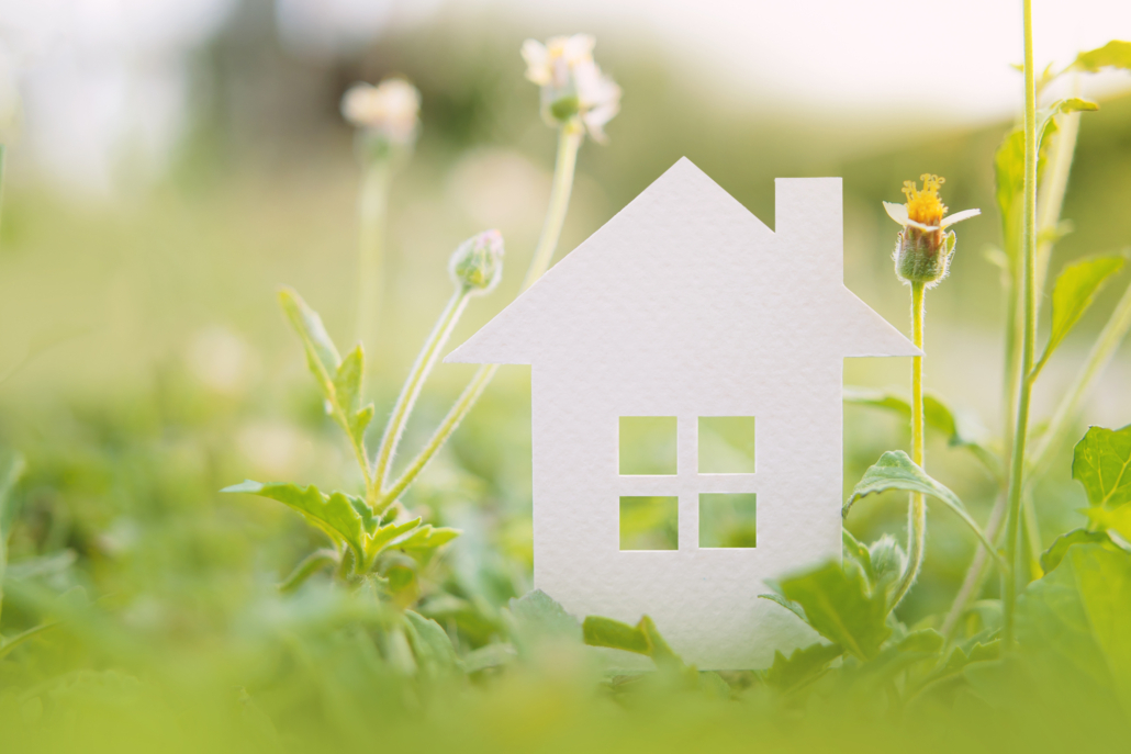 Papieren huis omringd door groen gras en bloemen, symbool voor duurzaam wonen en milieuvriendelijke maatregelen.