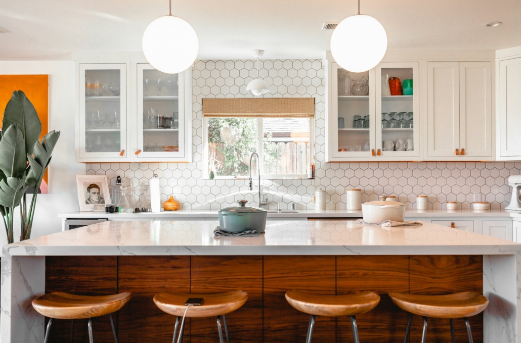 Een moderne keuken met een licht interieur, een groot marmeren kookeiland met houten krukken, witte kasten met glazen deuren en een honingraat tegelachterwand. Het daglicht valt door een groot raam met een bamboe rolgordijn, en er zijn decoratieve elementen zoals een groene plant en oranje accenten.